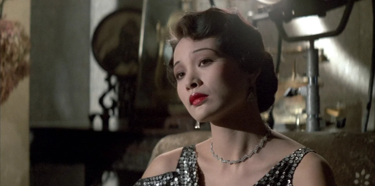 The Last Emperor (1987) movie still: closeup — A dimly lit room contains a vanity mirror and a professional studio light stand. A woman with dark…