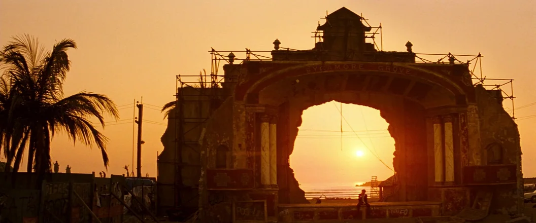 Romeo + Juliet (1996) movie still: wide — A decaying, elaborate stone archway stands on a beach covered in scaffolding structures. The sun sets…