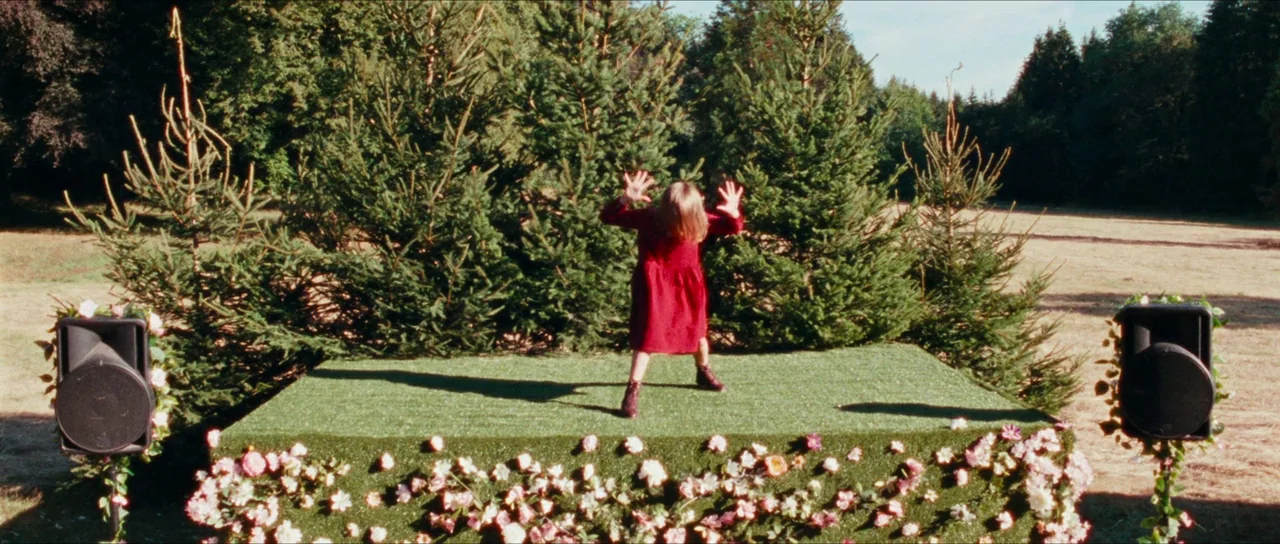 Inexorable (2021) movie still: wide — A square platform covered in artificial turf is placed in a sunny outdoor field. A young child in a red dress…