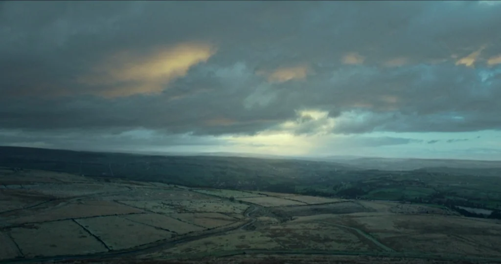 God’s Own Country (2017) movie still: establishing — A vast, rolling landscape of moorland features partitioned fields under a heavy, overcast sky.…