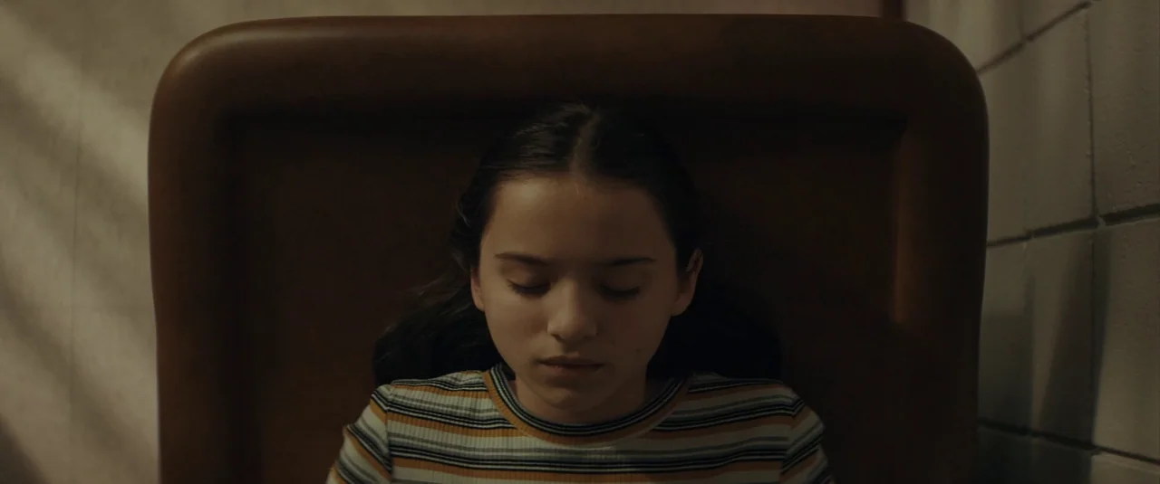 The Black Phone (2021) movie still: closeup — A girl rests against a high-backed tan leather chair in a room. Natural light filters in from the side.