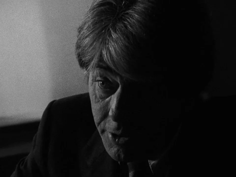 Following (1998) movie still: closeup — A dark, dimly lit indoor space with a man partially shrouded in shadow. The man focuses forward while…