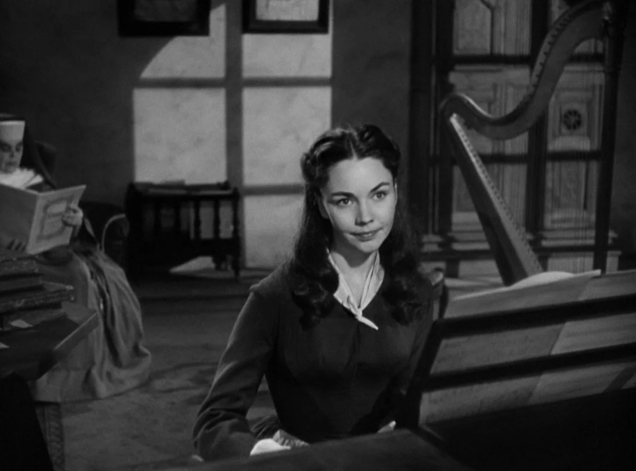 Madame Bovary (1949) movie still: medium — A dimly lit room contains a piano, a harp, and an seated nun reading. A woman in a dark dress plays the…