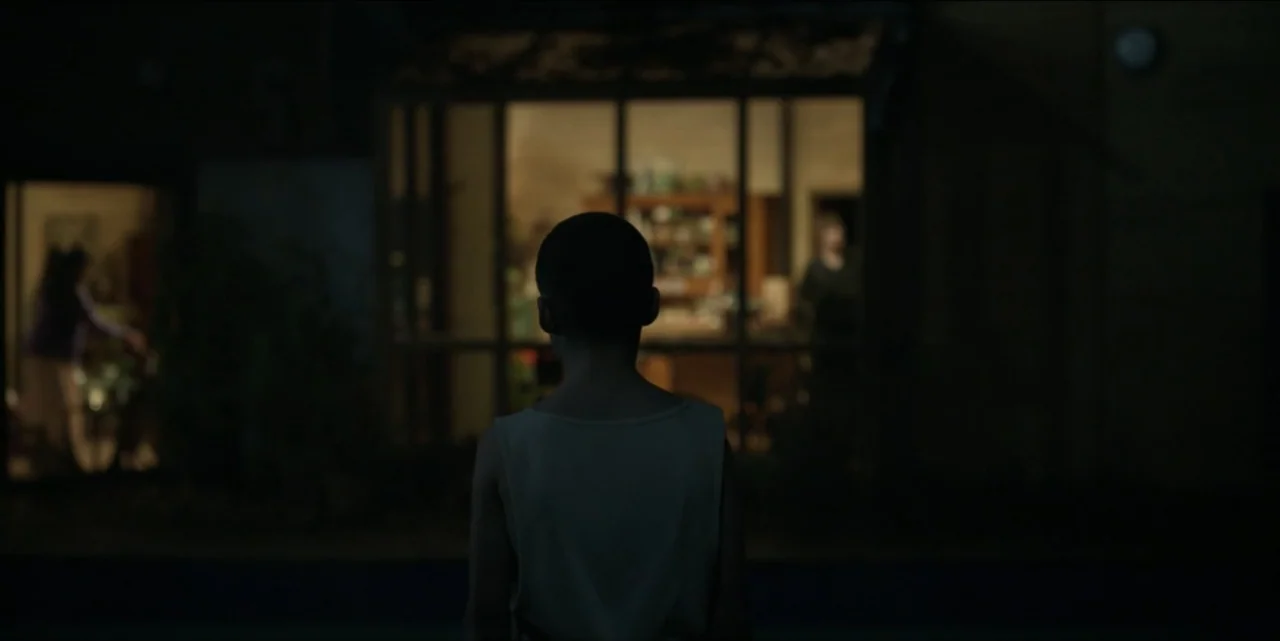 Bring Her Back (2025) movie still: medium — A dark outdoor space looks toward the brightly lit windows of a residential structure. A child stands in…