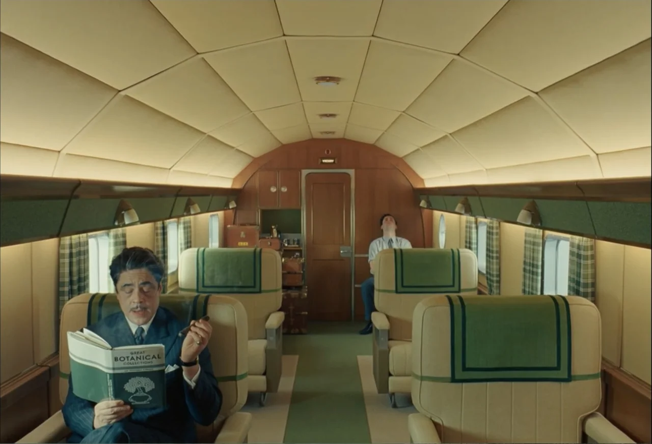 The Phoenician Scheme (2025) movie still: fullbody — A symmetrical view looking down the aisle of a vintage propeller airplane cabin. A man in a…