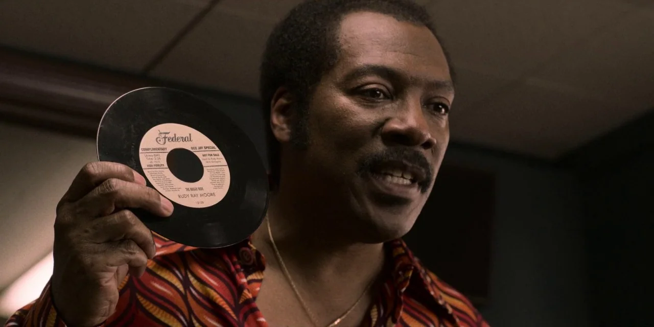 Dolemite Is My Name movie still: closeup — A dimly lit recording studio space with acoustic paneling visible in the background. A man holds up a…