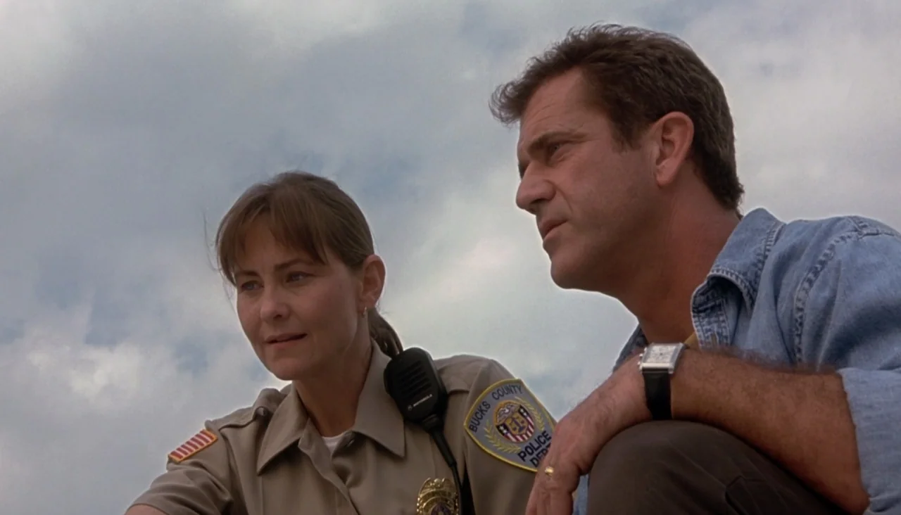 Signs (2002) movie still: medium — A woman in a police uniform and a man crouch together in an open field. The pair looks off-screen toward an…