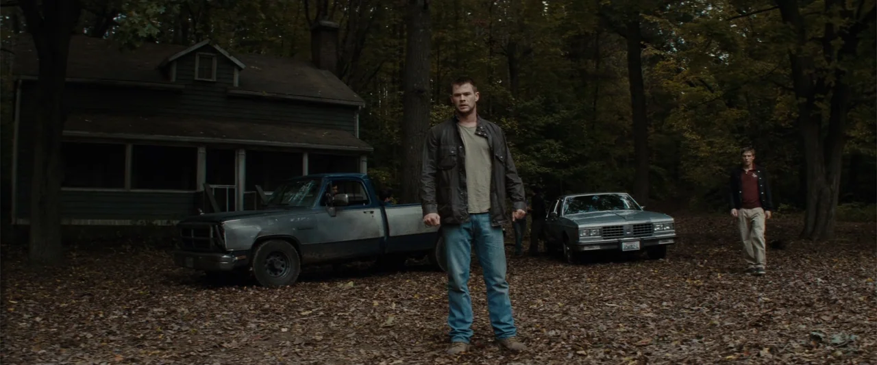 Red Dawn (2012) movie still: fullbody — A secluded dirt clearing in a dense forest features an old weathered house. A man stands in the foreground…