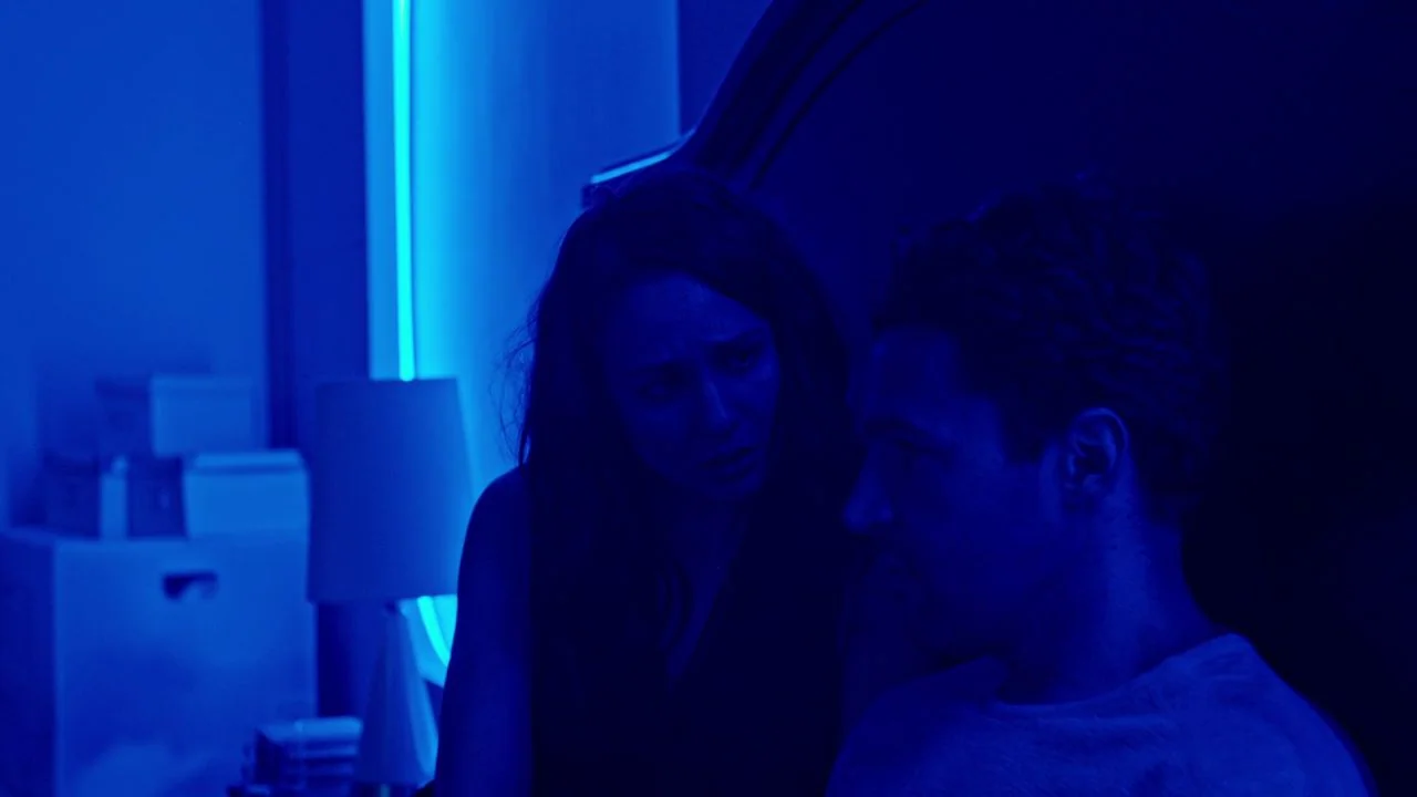 Possessor (2020) movie still: medium — A dimly lit room is illuminated by a sharp vertical blue neon light fixture. A man and a woman sit close to…