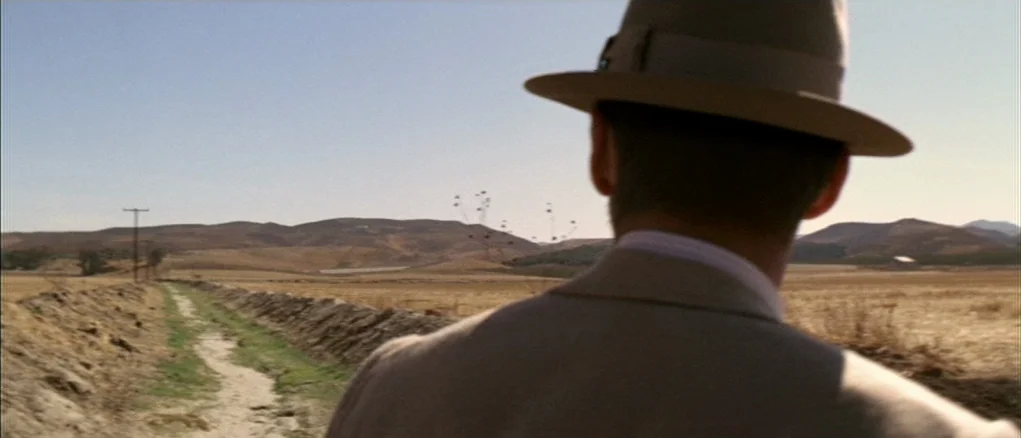 Chinatown (1974) movie still: medium — A vast, sun-drenched dry field stretches toward low rolling mountains under a clear sky. A man seen from…