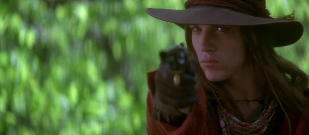 Ride With The Devil movie still: closeup — A dense patch of blurred green foliage serves as the backdrop. A person in period clothing points a…