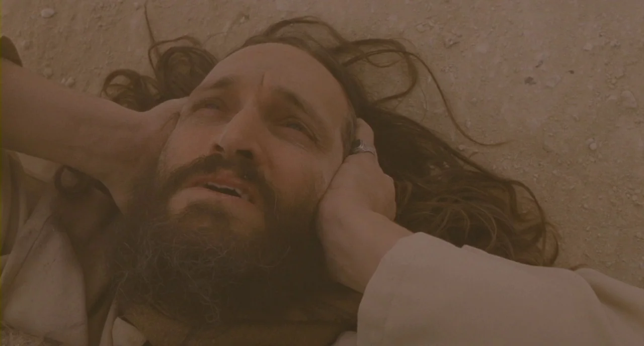 Essential Killing (2010) movie still: closeup — A man lies prone on an expansive, featureless stretch of arid desert sand. He holds his hands…