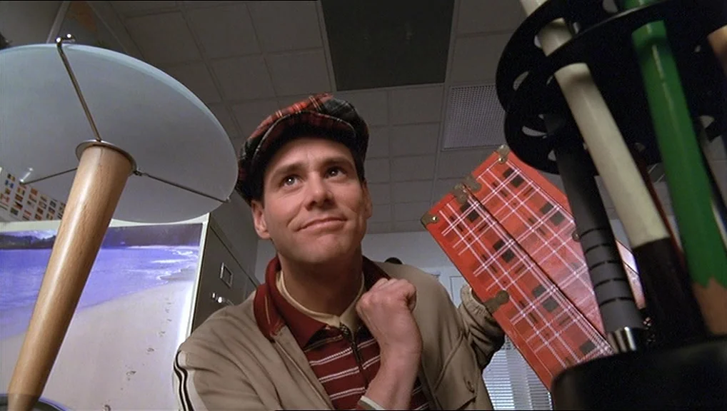 The Truman Show movie still: medium — A travel agency interior features a poster of a beach on the wall. A man wears a plaid hat and holds a red…