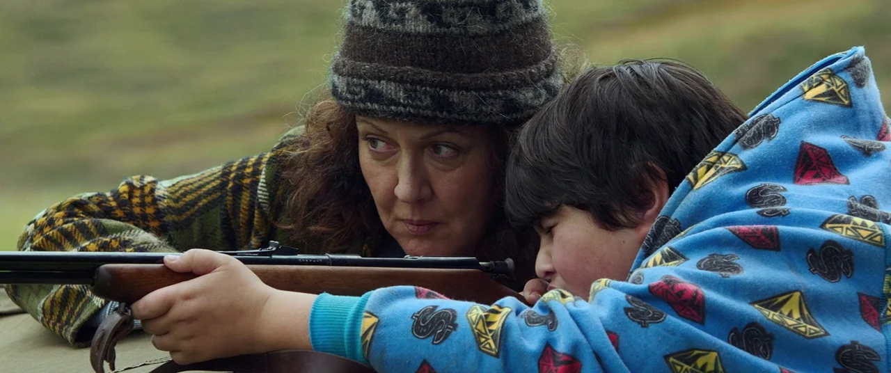 Hunt for the Wilderpeople (2016) movie still: medium — An adult woman and a young boy crouch in an outdoor grassy area. They share the aim of a…