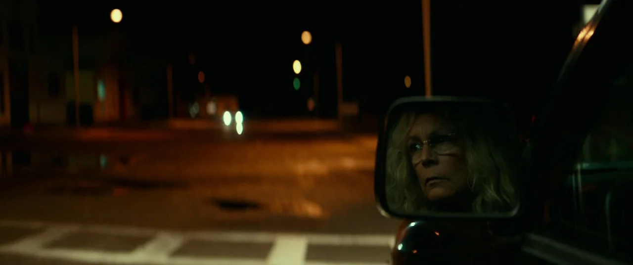 Halloween (2018) (2018) movie still: wide — A dark, empty intersection at night with distant bokeh streetlights glowing in the distance. A woman's…