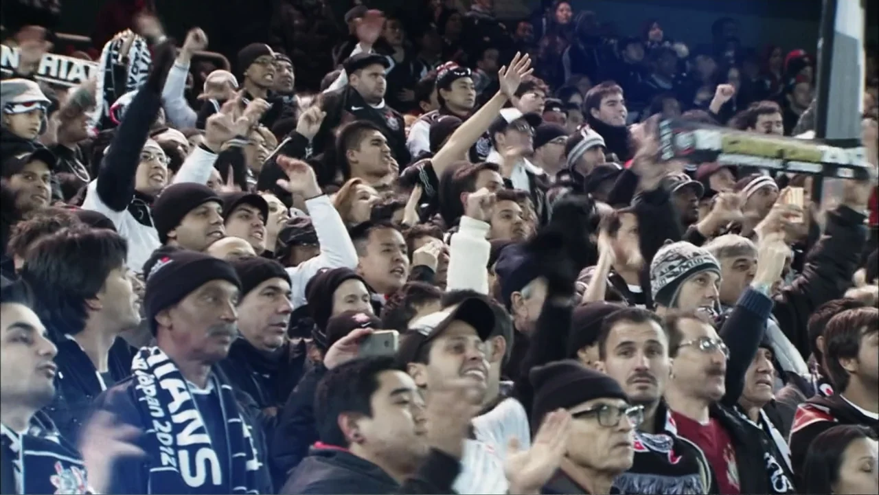 Vai Corinthians (2023) movie still: wide — A dense crowd of spectators sits in stadium bleachers during an evening sporting event. People are…