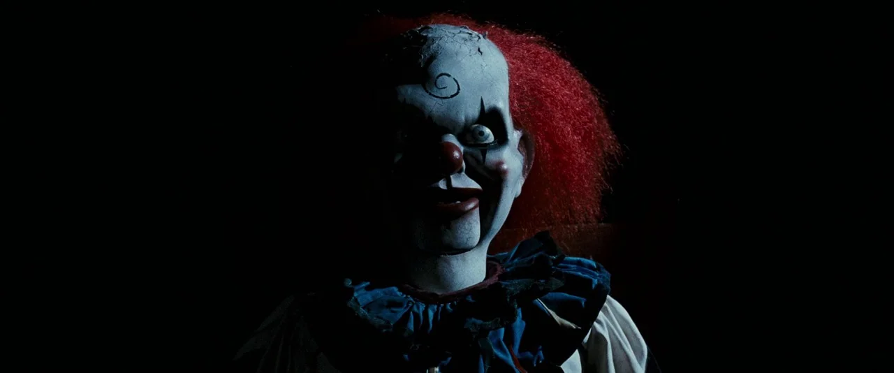 Dead Silence (2007) movie still: closeup — A clown-faced ventriloquist dummy appears against an pitch black void. The puppet stares forward with…