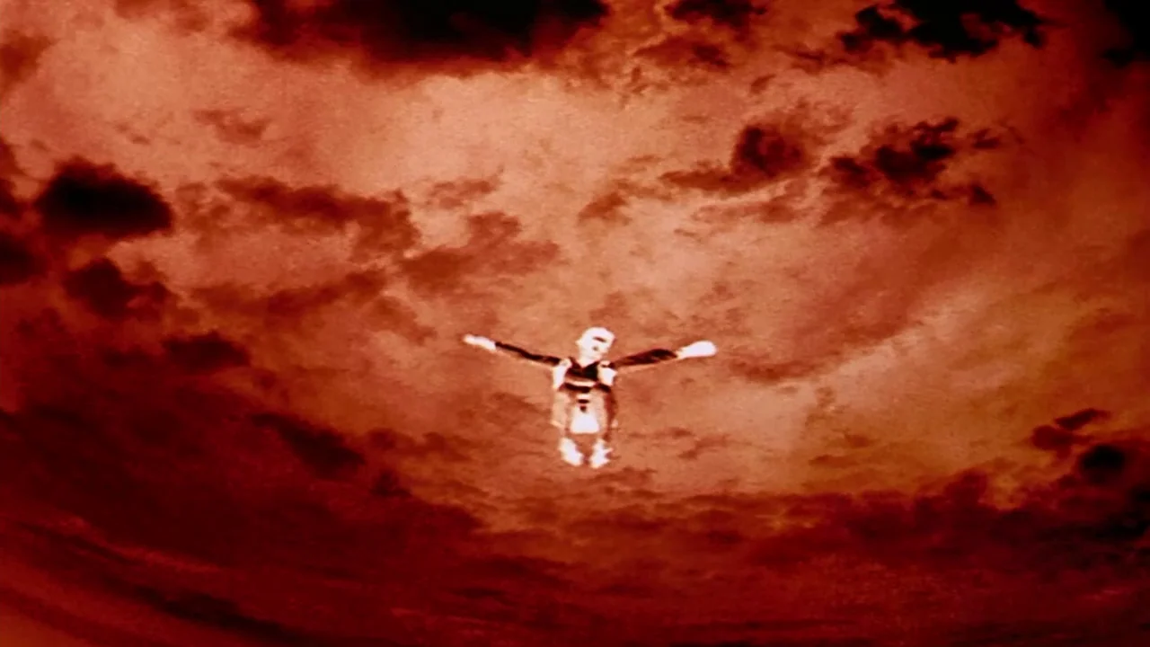 Naqoyquatsi (2002) movie still: wide — A person falls through a dark red, cloud-filled sky. The individual is positioned centrally, limbs extended…