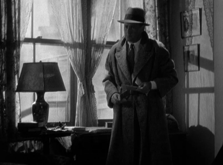 Blonde Venus (1932) movie still: medium — A dark apartment room features lace-covered windows and framed pictures on the wall. A man in a trench…