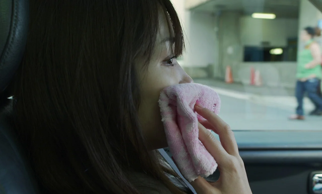 Like Someone In Love (2012) movie still: closeup — Inside a vehicle, the view through the passenger side window shows an outdoor area. A young woman…