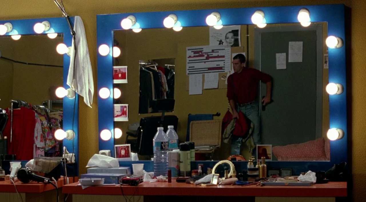 Tie Me Up! Tie Me Down! (1989) movie still: medium — A cluttered backstage room with yellow walls and two large blue vanity mirrors. A man in a red…
