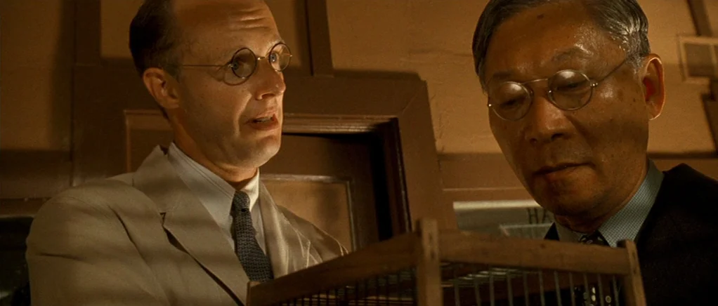 King of the Hill (1993) movie still: medium — A dimly lit room with plain walls contains a wooden cage on a table. Two men in formal 1940s attire…