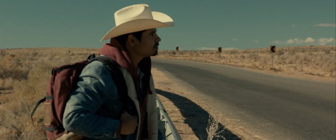 Frontera (2014) movie still: medium — An arid highway stretch in a dry desert landscape during bright daylight conditions. A man in a cowboy hat and…