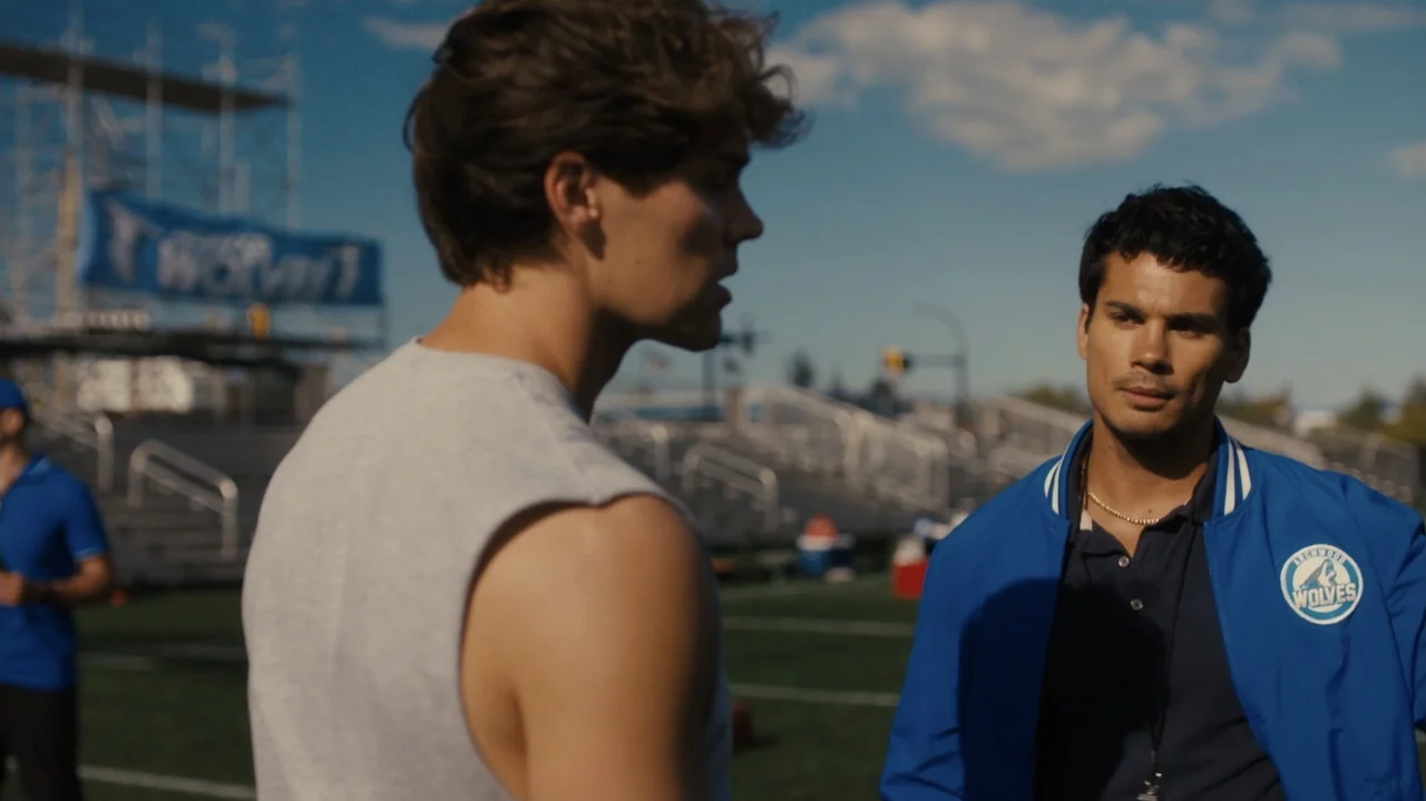 Sidelined: The QB and Me (2025) movie still: medium — A sports field with metal bleachers and a blue team banner in the background. Two young men…