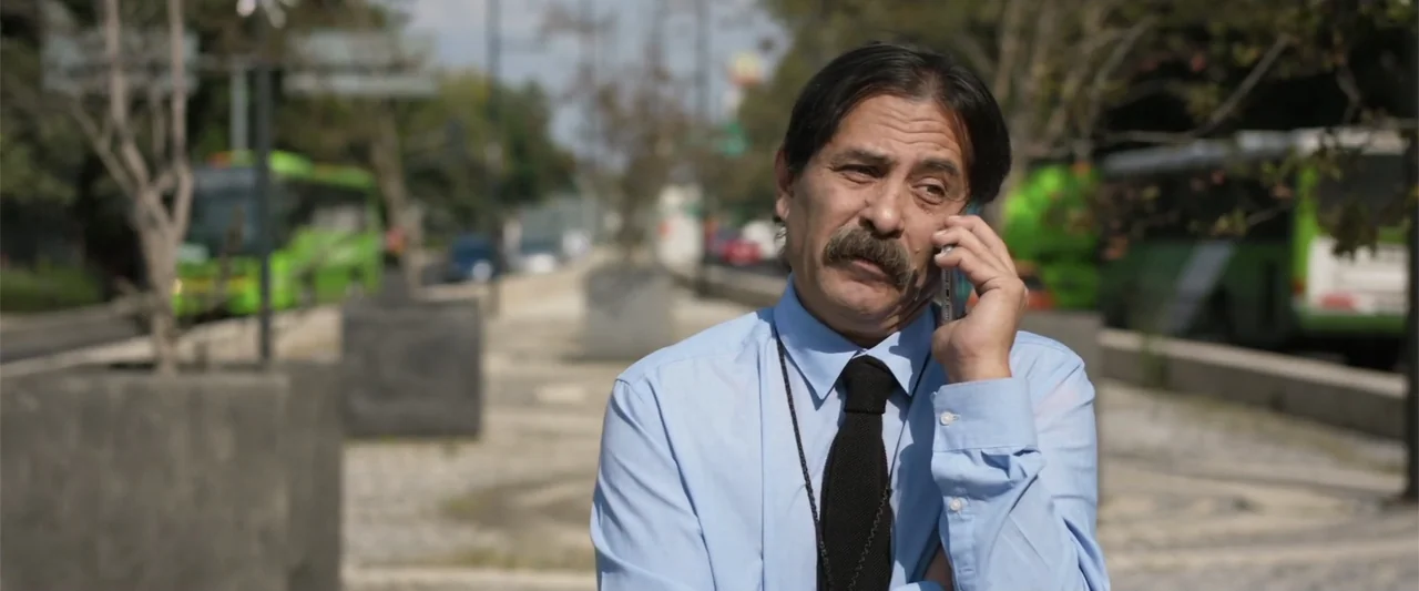 Winner Takes the Cake (2025) movie still: medium — A sidewalk sits alongside a busy urban thoroughfare with a green bus in background. A middle-aged…