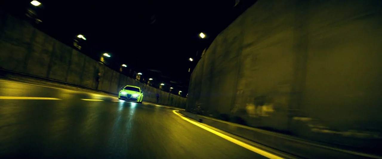 6 Underground (2019) movie still: wide — A long, curved concrete tunnel at night illuminated by rows of overhead fluorescent lights. A bright yellow…
