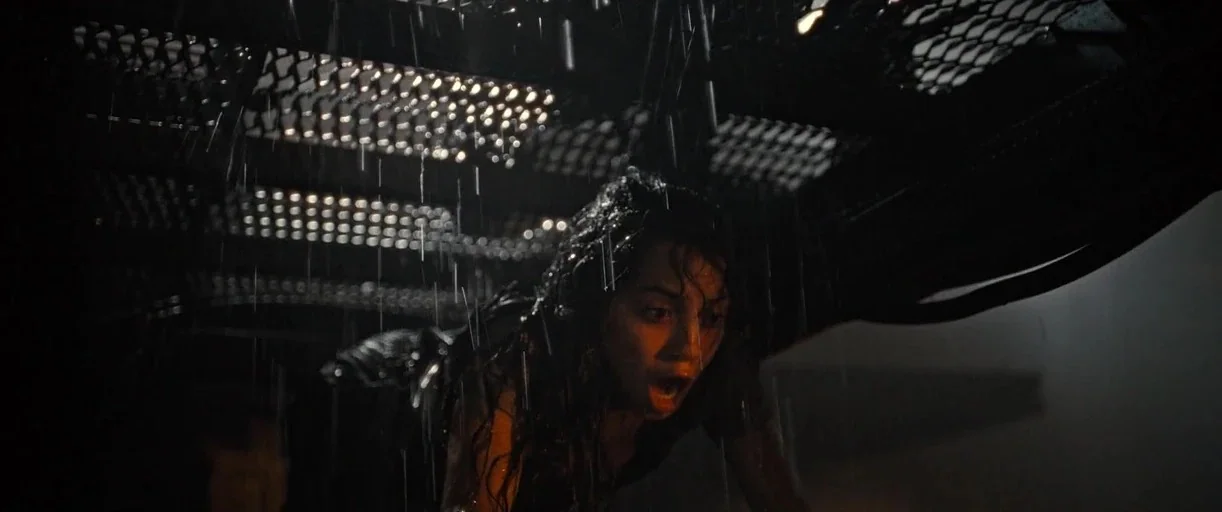 Alien: Romulus (2024) movie still: closeup — A woman stands under a dark metal grate structure during a heavy rainstorm at night. She looks forward…