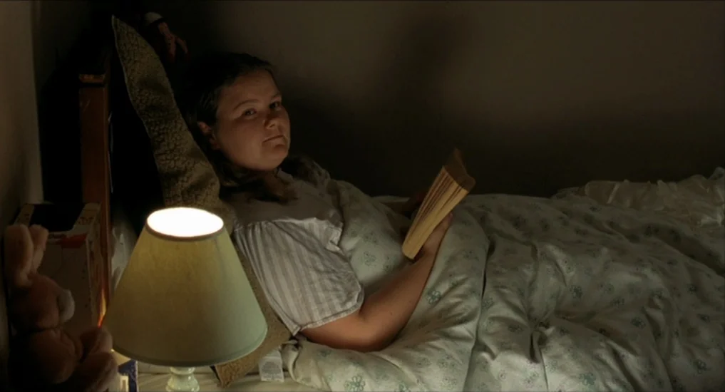 All or Nothing (2002) movie still: medium — A dimly lit room with a subject lying under floral patterned bedding. The subject is propped against…