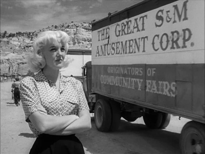 Ace In The Hole (1951) movie still: medium — An open dirt lot features a parked amusement truck with large painted lettering. A blonde woman with…