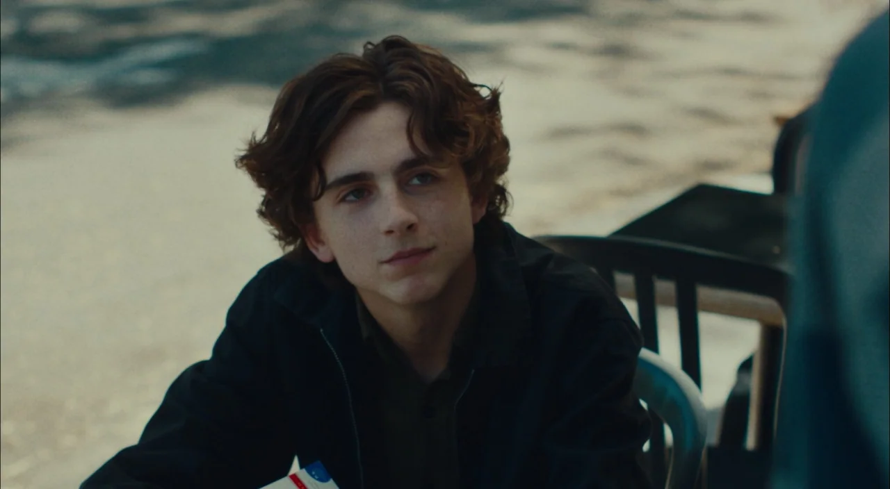 Lady Bird (2017) movie still: medium — A young man sits at an outdoor table with a textured, blurred surface behind him. He tilts his head slightly…