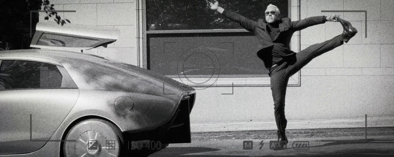 The Naked Gun (2025) movie still: fullbody — A man balances on one leg beside a parked vehicle in front of a building. The composition includes a…