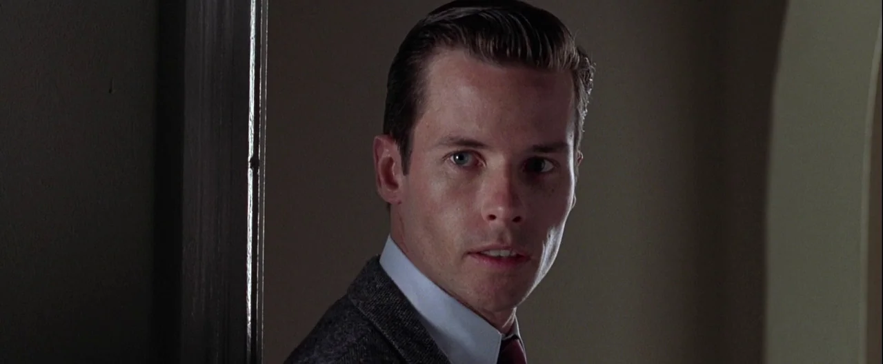 L.A. Confidential (1997) movie still: closeup — A man stands near a dark wooden door frame in a dimly lit room. He looks toward the side of the…