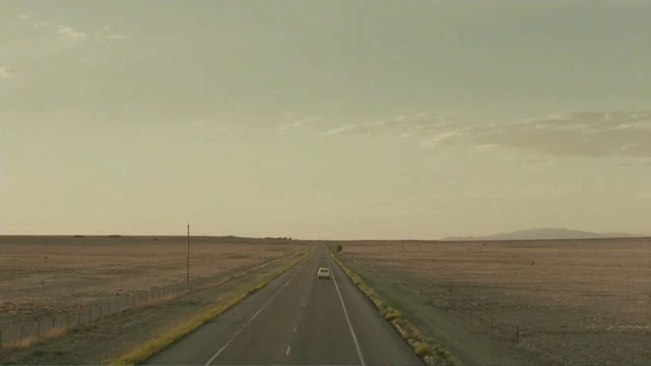Legion (2010) movie still: establishing — An empty two-lane asphalt road stretches into a flat, dry desert landscape at sunset. A single white car…