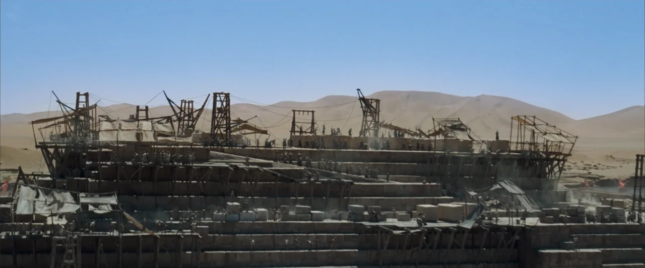 10,000 BC (2008) movie still: wide — An ancient pyramid under construction sits amidst vast sand dunes under a clear sky. Hundreds of laborers work…