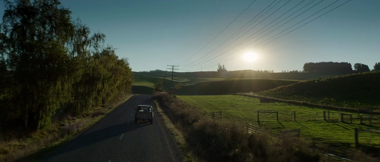 Pete’s Dragon (2016) movie still: wide — A rural paved road winds through rolling green hills under a clear bright sky. Utility lines stretch across…