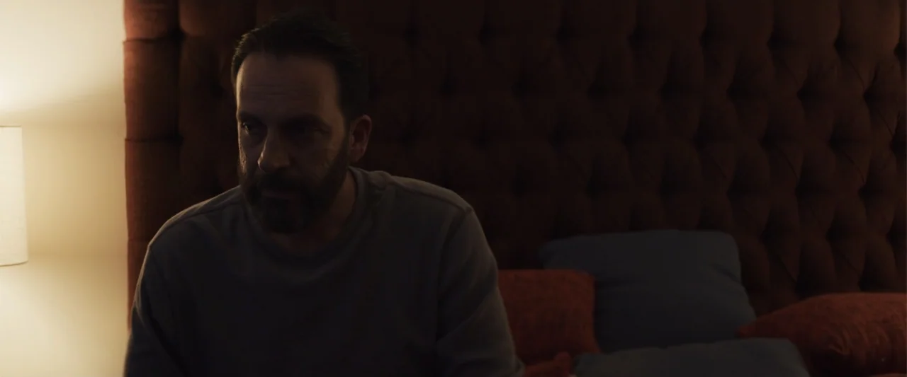 Human Animals (2020) movie still: medium — A dark bedroom features a large tufted velvet headboard against the wall. A man sits upright in bed…