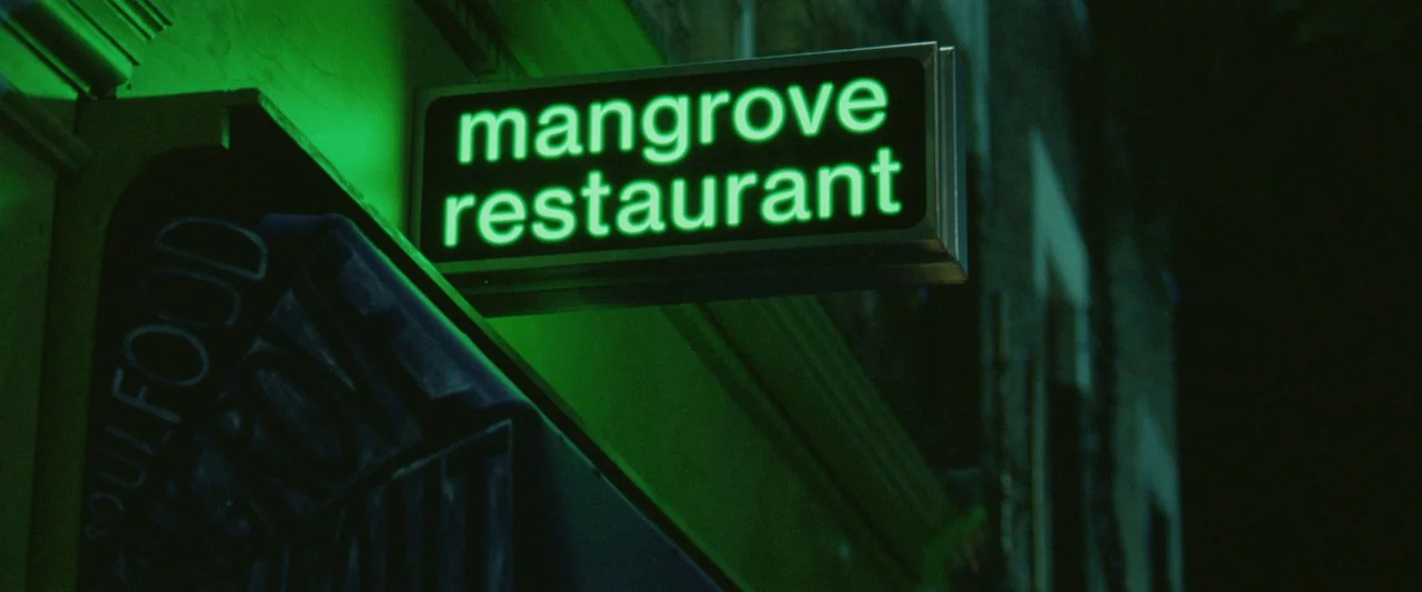 Mangrove (2020) movie still: closeup — A night exterior shot features a glowing green sign hanging above a dark storefront. The sign displays the…