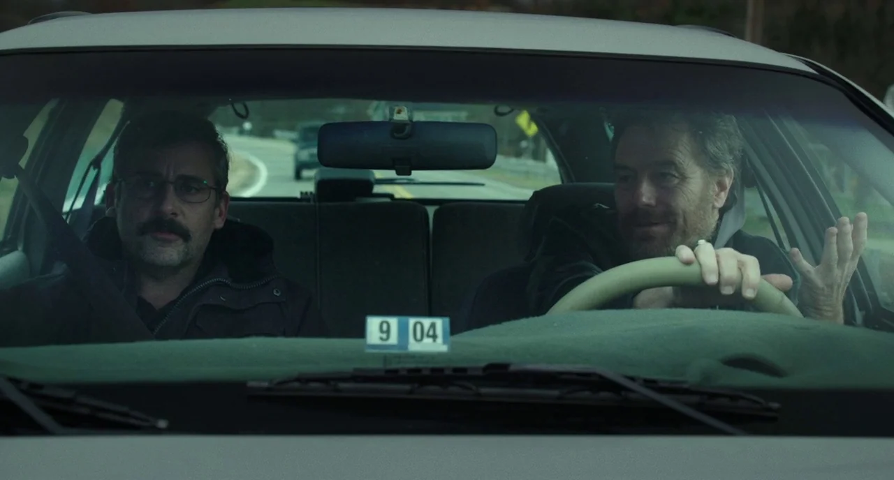Last Flag Flying (2017) movie still: medium — Two men occupy the front seats of a sedan during a daytime drive. The driver keeps his hand on the…