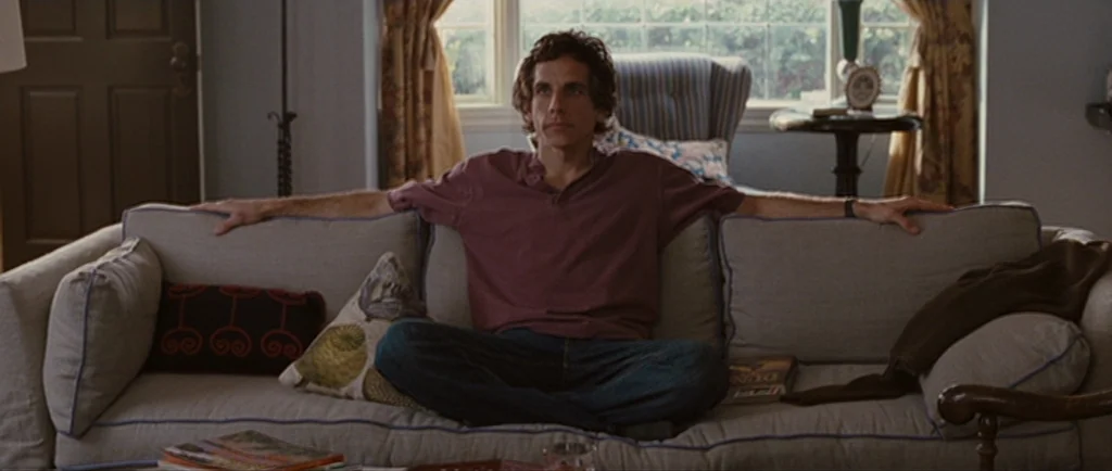 Greenberg (2010) movie still: medium — A well-lit domestic space contains a beige sofa positioned in front of a window. A man sits in the middle of…