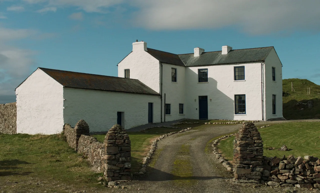 It Is In Us All (2022) movie still: establishing — A two-story whitewashed farmhouse with a slate roof stands at the end of a path. A dry-stone wall…