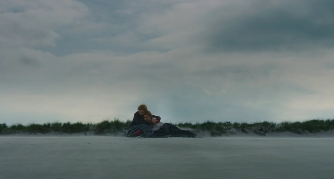 Little Women (2019) movie still: wide — A vast, flat beach under a heavy grey sky with sparse dune grass. Two women are sitting together on the…