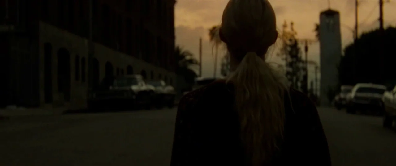 Low Down (2014) movie still: medium — A city street at golden hour with parked cars lining both sides. A young woman with a blonde ponytail walks…