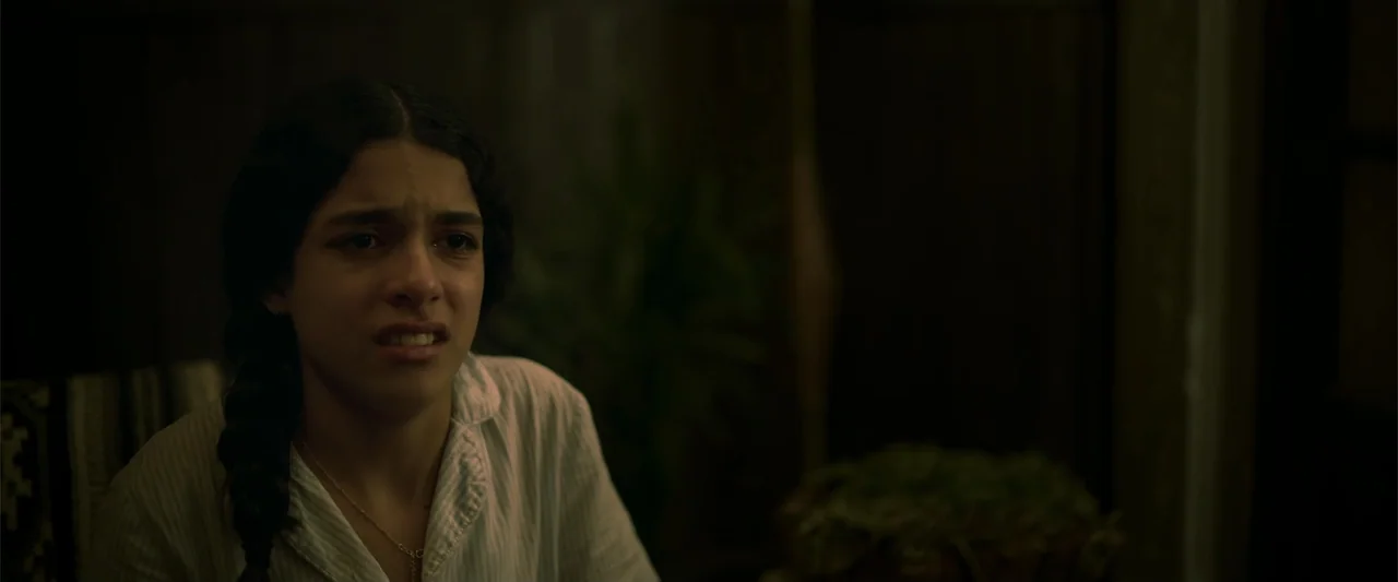 Evil Eye (2020) movie still: medium — A dimly lit room with dark wooden wall textures in the background. A young woman with a braid sits in a white…