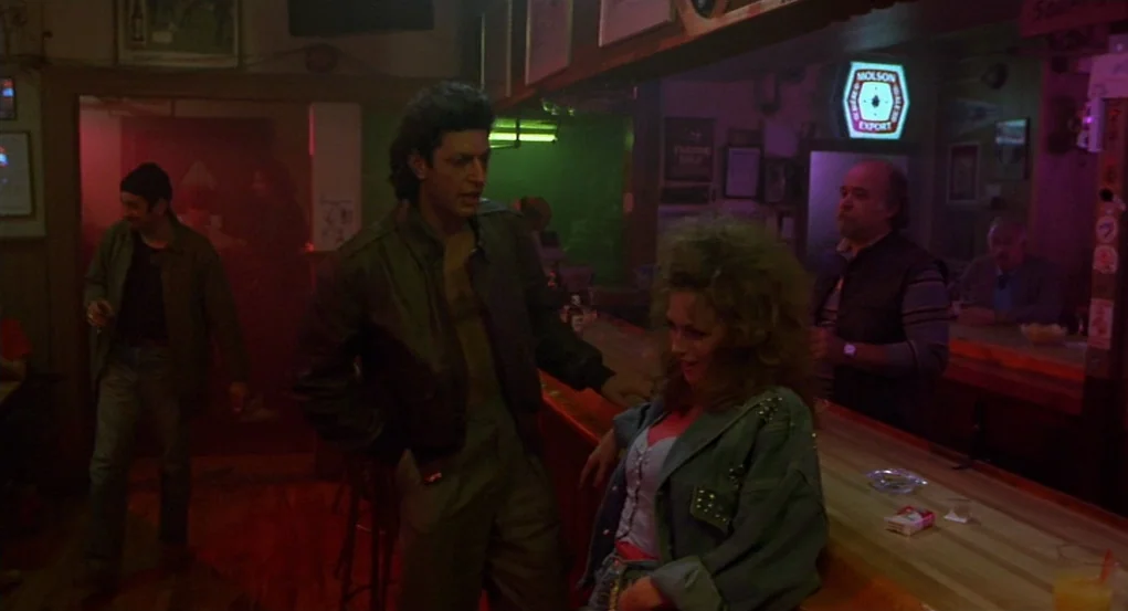 The Fly (1986) movie still: medium — A dimly lit bar with neon signage and dark wooden surfaces. A man and woman stand at the counter while others…