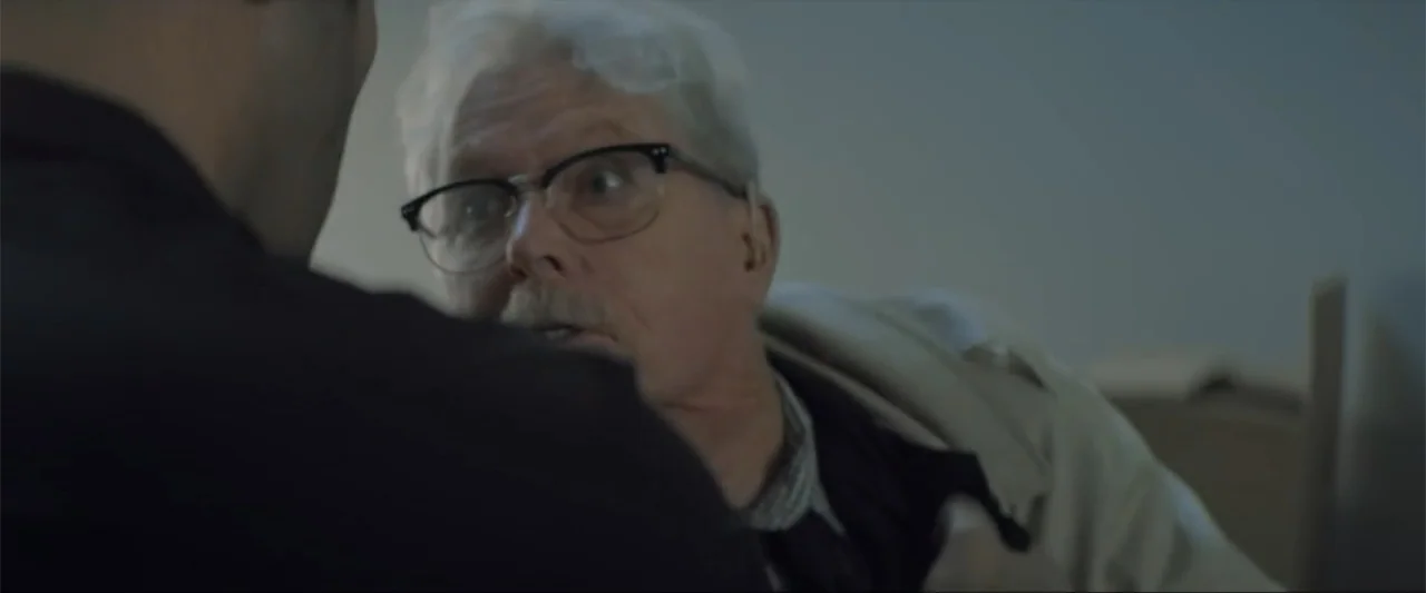 Gender Under Attack (2018) movie still: medium — A dimly lit interior space with neutral walls and soft shadows. An older man with white hair and…