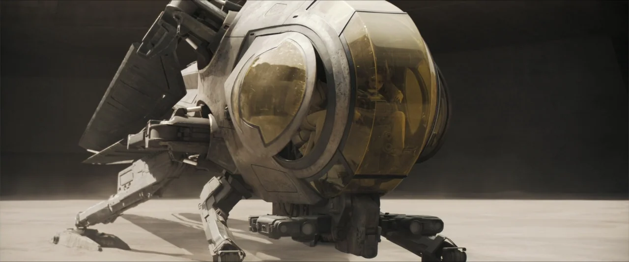 Dune (2021) movie still: medium — A futuristic aircraft with a yellow canopy sits on a concrete hangar floor. A male pilot wearing a light flight…